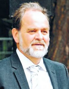 Ein leicht seitliches Portrait von Dr. Hans-Uwe Feige. Er trägt ein weißes Hemd mit einem dunkelgrauen Sakko und gestreifter Krawatte.