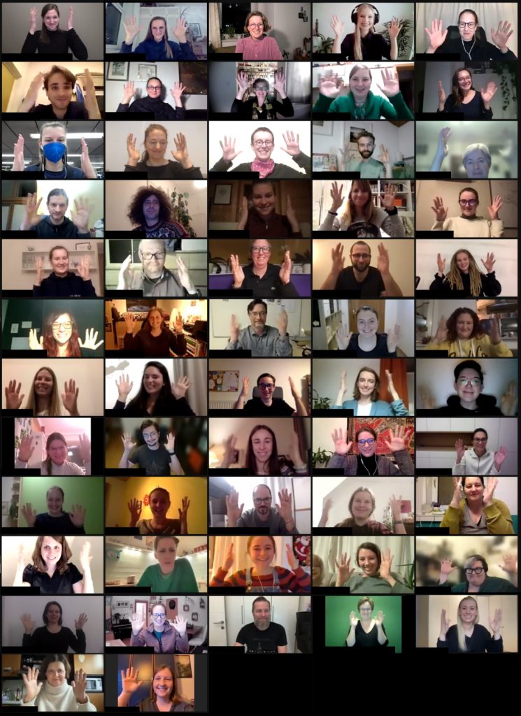 Screenshot der Zoom-Videokonferenz. 57 kleine Fenster zeigen Teilnehmer*innen, die lächeln und ihre Hände schütteln, um zu applaudieren.