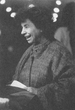 Schwarz-weiß Fotografie, die Ursula Bellugi zeigt. Frau mit kurzem dunklen Haar, das wild nach oben steht, lachend, schlicht gekleidet