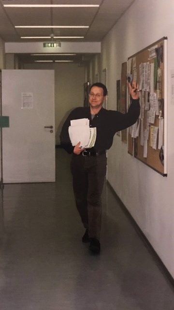 Das Foto zeigt den lächelnden Jens Heßmann beim Entlanggehen eines Flures in einem Gebäu-de der Hochschule Magdeburg-Stendal. In seiner rechten Hand trägt er einen Stapel weißes Pa-pier und die linke Hand hebt er winkend zum Gruß.